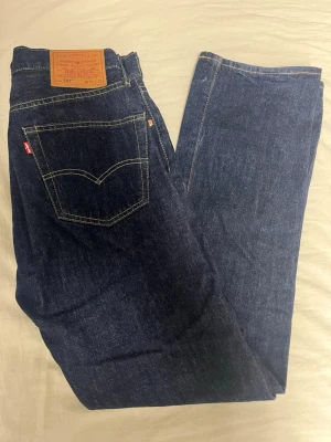 Levi's 501 mörkblå jeans - Klassiska Levi's 501 jeans i mörkblå tvätt med raka ben och normal passform. Jeansen har fem fickor, synliga sömmar och den ikoniska läderpatchen bak i midjan. Tillverkade i slitstarkt bomullsjeansmaterial och har knappgylf framtill. Använda få gånger nästan som nya