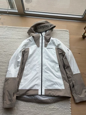 Peak Performance jacka XL beige/vit - Snygg jacka från Peak Performance i storlek XL. Färgen är en fräsch kombo av beige och vit med svart Peak Performance-logga på bröstet och huvan. Jackan har huva, hel dragkedja och är tillverkad i ett slitstarkt syntetmaterial som passar perfekt för utomhusaktiviteter.