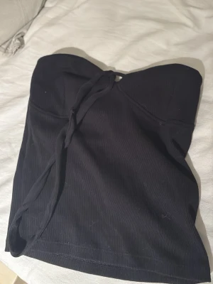 Svart ribbad tubtopp med snörning - Säljer en svart ribbad tubtopp med snygg snörning framtill. Toppen är axelbandslös och har en croppad passform som passar perfekt till sommarens alla outfits. Materialet är mjukt och stretchigt, vilket gör den bekväm att bära hela dagen. 