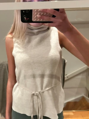 Beige ärmlös polotopp med knyt - Säljer en beige polotopp i mjukt material med ärmlös design och ett snyggt knyt i midjan. Toppen har en hög krage som ger en cool look och är perfekt att styla med jeans eller kjol. Bältet är justerbart!! 