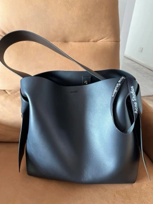 Svart axelväska från Acne Studios - Säljer en stilren svart axelväska från Acne Studios i slätt skinn. Väskan har en minimalistisk design med bred axelrem och en snygg detalj med Acne Studios-band. Perfekt för dig som gillar enkel och modern stil. nypris 18.000kr
