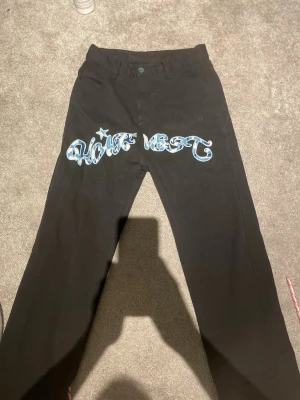 Svarta byxor med blå camo-tryck - Svarta byxor med unika blå camo-tryck och stjärnor på både fram- och baksidan. Framsidan har ett stort graffiti-inspirerat tryck med text och stjärna, medan baksidan har två stora stjärnor med blått camouflagemönster. Raka ben och klassisk midja, tillverkade i bomull.