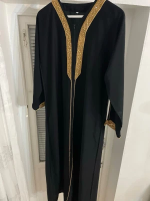 Svart långklänning med guldbroderi - Svart långklänning med breda ärmar och detaljerad guldbroderad kant längs halsringningen och ärmsluten. Klänningen har en slits på sidan och stängs med dragkedja framtill. Materialet känns lätt och faller snyggt. Perfekt för dig som gillar eleganta och unika plagg. Klänningen är 135cm lång och är storlek M men passar även S och XS. Använd en gång