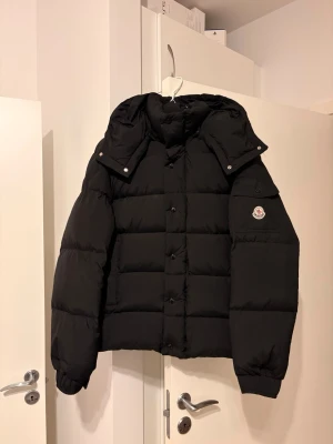 Moncler Vezere I M - Säljer en svart pufferjacka från Moncler med avtagbar huva och klassisk logga på ärmen där jackan inte passar mig och jag kan tyvärr inte lämna tillbaka den. Jackan har quiltad design, knappar och dragkedja framtill samt elastiska muddar vid ärmsluten. Perfekt för kalla dagar och riktigt snygg. 