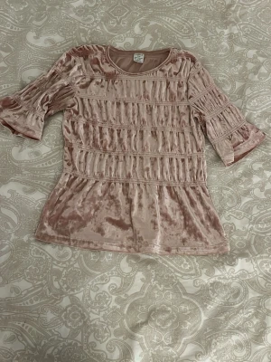 Rosa sammetsblus från Lindex - Supersöt rosa sammetsblus från Lindex med rynkade detaljer och halvlånga trumpetärmar. Blusen har en mjuk glansig yta och rund halsringning. Perfekt för dig som vill ha en unik och trendig topp med lite extra volym i ärmarna.