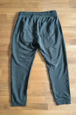 Mörkgrå mjukisbyxor med snörning - Säljer ett par mörkgrå mjukisbyxor med ribbade muddar vid benslut och midja. Byxorna har snörning i midjan, två sidofickor fram och en bakficka. Perfekta för chill eller träning, tillverkade i mjukt bomullsmaterial.