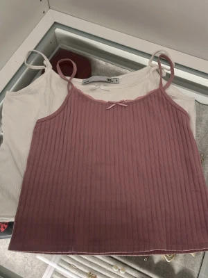 Ribbad rosa och vit linnetopp lager 157 och Zara - Två stilrena linnetoppar från lager 157 och Zara i storlek S. Ena toppen är ljusrosa med ribbad struktur och en liten rosett framtill, den andra är vit och slät. Båda har smala axelband och är perfekta för varma dagar.