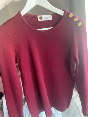 Vinröd stickad tröja från Busnel - Snygg vinröd stickad tröja från Busnel med rund hals och guldknappar med B på axeln. Tröjan har lång ärm och ett litet Busnel-emblem vid mudden. Nypris 3200kr! Storlek 40, väldigt liten i storleken!