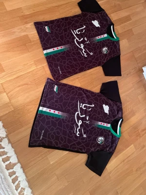 Syrien tröja  - Svart sporttröja med den nya Syrien-flaggan upp och ner på framsidan. Perfekt för träning, vardag eller som ett unikt statement-plagg. Skön och slitstark, med en sportig passfor. Både för kvinnor och män