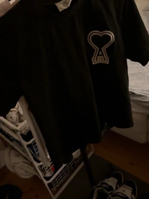 Svart t-shirt från Ami  - Svart t-shirt från Ami med korta ärmar och en stor broderad hjärtdetalj med bokstaven A på bröstet. Klassisk rund hals och tillverkad i mjuk bomull. Perfekt för dig som gillar stilrena plagg med en twist. storlek xs eller s