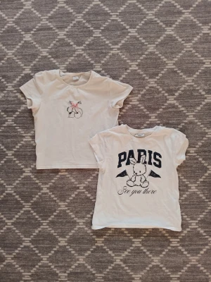Två vita t-shirts med tryck, Lindex & Kappahl - Säljer två vita t-shirts i bomull, en från Lindex med körsbär och rosett i rosa, och en från Kappahl med Paris-tryck och en söt kanin.🍒🐇Båda har rund hals och korta ärmar, perfekt för dig som gillar gulliga motiv och mjuka material. Båda för 30 kr+frakt