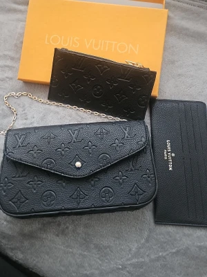 Svart handväska  - Säljer en svart handväska med klassiskt monogrammönster i skinn. Väskan har en snygg kedja som axelrem och medföljande plånbok och korthållare i samma stil. Perfekt för dig som gillar lyxiga detaljer och ikonisk design.