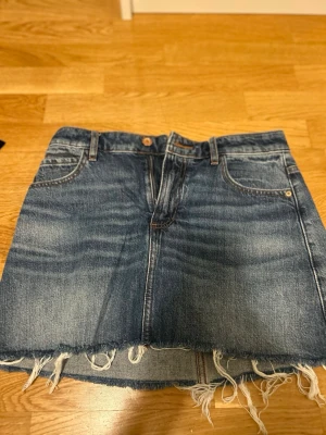 Blå jeanskjol från Zara, strl S - Säljer en blå jeanskjol från Zara i storlek S med klassisk femficksdesign och rå, fransig kant nedtill. Kjolen har dragkedja och knapp framtill samt bälteshällor. Perfekt för dig som gillar en avslappnad och trendig stil.