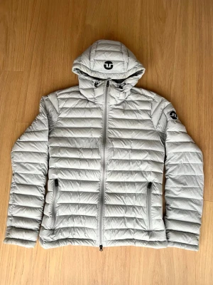 Ljusgrå dunjacka från J.Lindeberg - Säljer en stilren ljusgrå dunjacka från J.Lindeberg med huva och dragkedja framtill. Tunn dunjacka som fungerar lika bra med jeans som vaddering under en skaljacka. Storlek L men sitter tight som en M. 