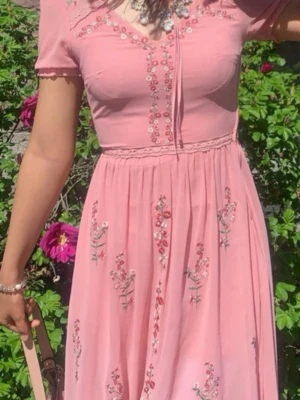 Rosa långklänning med broderade blommor - Superfin rosa långklänning med broderade blommor i rött och vitt. Klänningen har korta ärmar, v-ringad hals och dekorativ knytning framtill. Materialet känns lätt och luftigt, perfekt för varma dagar. Söt spetsdetalj vid midjan och romantiska broderier ger en extra charm. Xs / s