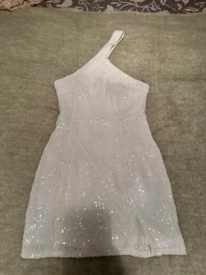 Glittrig vit one shoulder klänning - Säljer en snygg vit kortklänning från Nelly x Glamorous med glittriga paljetter över hela tyget. Klänningen har en trendig one shoulder-design och en liten slits framtill. Perfekt för dig som vill sticka ut med en unik look.