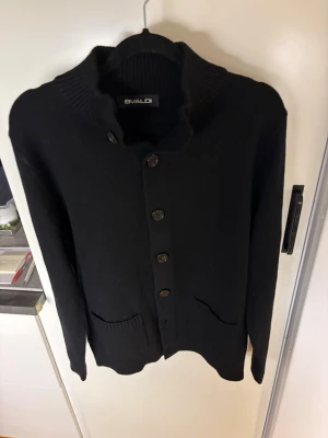 Svart kofta från HELT NY! - Svart kofta  med ribbad krage och fyra knappar framtill. Koftan har två fickor och är långärmad, perfekt för lager-på-lager. Materialet är stickat och ger en mjuk känsla. Enkel och stilren design som funkar till många olika looks. Den har aldrig använts bara testat så inga skador. 