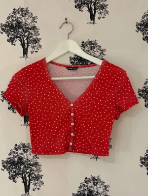Röd croppad topp med hjärtan från Shein - Säljer en röd croppad topp från Shein med små vita hjärtan som mönster. Toppen har korta ärmar, v-ringning och knappar framtill. Perfekt för dig som gillar söta detaljer och vill ha en färgglad look.