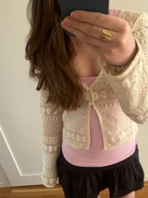 Beige stickad kofta med spetsdetaljer - Säljer en beige stickad kofta med spetsdetaljer och dekorativa mönster. Koftan har lång ärm och knäpps framtill med små knappar. Perfekt att slänga över en topp för en söt och trendig look Endast använd en gång.