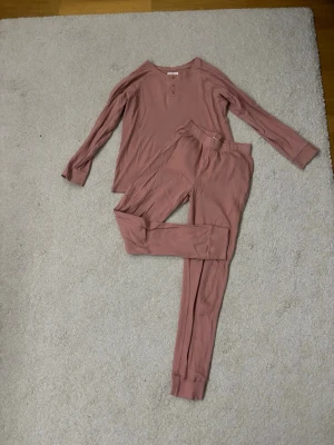 Rosa ribbat långärmat pyjamasset i ekologisk bomull - Säljer ett mjukt och ribbat pyjamasset i en snygg rosa färg från organic cotton. Långärmaden har rund hals, tre knappar framtill och är gjord i ekologisk bomull. Perfekt för dig som gillar enkel och chill stil. Skicka för fler bilder!