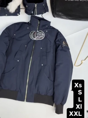 Moose Knuckles bomberjacka med dragkedja - Snygg bomberjacka från Moose Knuckles i mörkblått och svart. Jackan har hög krage, ribbade muddar och flera fickor med tryckknappar. Metallmärke på ärmen och grov dragkedja framtill. Perfekt för kalla dagar.Helt nya oanvänd 