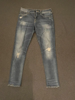 True Religion blå jeans med slitningar - Säljer ett par blå jeans från True Religion i storlek 32. Modellen har slim passform och är dekorerad med slitningar och snygga detaljer på benen. Klassisk femficksdesign, gul kontrastsöm och broderade bakfickor. Materialet är jeans med en skön känsla.