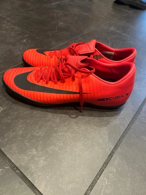 Nike MercurialX röda fotbollsskor - Säljer ett par Nike MercurialX fotbollsskor i röd färg med svarta detaljer och snörning. Skorna har en låg profil och platt sula med mönstrad undersida för bra grepp på planen. Materialet är syntet och designen har tunna svarta ränder längs ovansidan.