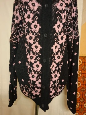 Svart kofta med rosa blommor och prickar - Svart stickad kofta med knappar framtill, dekorerad med rosa blommor på framsidan och rosa prickar på baksidan och ärmarna. Koftan har långärmad passform och ribbade muddar vid nederkant och ärmslut. Perfekt för dig som gillar coola mönster och mjuka material. Storlek 12 = M men eftersom jag provade den så säger jag Large, för jag är storlek Medium och den är för stor för mig. Made in England