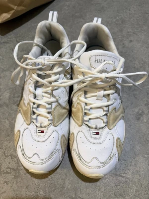 Tommy Hilfiger vita sneakers med mockadetaljer - Säljer ett par vita sneakers från Tommy Hilfiger med beige mockadetaljer och klassisk logga på tungan och sidan. Skorna har snörning, rund tå och platt sula. Materialmix av skinn och mocka ger en schysst kontrast och sportig vibe.