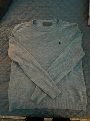 Grå merinoulltröja från Morris - Snygg grå rundhalsad tröja från Morris i 100% merinoull. Tröjan har ett diskret svart broderat emblem på bröstet och ribbade muddar vid ärmslut och nederkant. Perfekt för dig som gillar stilrena och klassiska plagg.