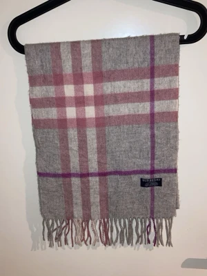 Grå och rosa rutig Burberry halsduk - Säljer en snygg halsduk från Burberry i klassiskt rutigt mönster med grå, rosa och lila toner. Halsduken är tillverkad i 100% lammull och har fransar längst ner.