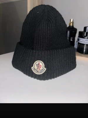 Svart Moncler mössa i ull - Snygg svart mössa från Moncler med klassisk ribbstickad design och uppvikt kant. Mössan har Moncler-logga framtill och är tillverkad i mjuk ull som håller dig varm. Perfekt för dig som vill ha en stilren accessoar med exklusiv känsla.