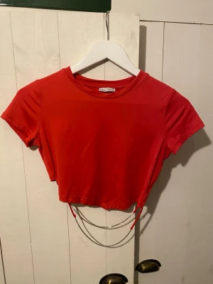 Röd croppad topp med diamantdetalj - Säljer en röd croppad topp från Bershka i mjuk bomull. Toppen har korta ärmar och coola kedjor med diamanter som hänger baktill. Tröjan är croppad och får en unik design av diamant hängena på ryggen. Perfekt för dig som vill sticka ut med en enkel men snygg detalj. Passar dig som gillar trendiga och unika plagg.