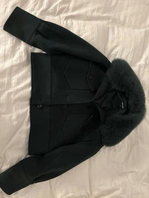 Svart bomberjacka med päls från Meotine - Säljer en svart bomberjacka från Meotine med stor fluffig päls på kragen. Jackan har två stora fickor med knappar framtill och är croppad i modellen. Materialet är mjukt och har en snygg satinliknande insida. Perfekt för dig som vill ha en cool och trendig look.