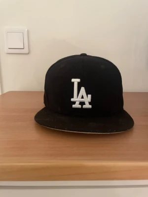 Svart LA Dodgers keps från New Era - Säljer en svart LA Dodgers snapback från New Era med vit broderad LA-logga framtill och 1988 World Series-märke på sidan. Kepsen har platt skärm och broderade MLB-detaljer bak. Perfekt för dig som gillar sportig streetstyle.