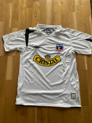 Colo-Colo matchtröja från Umbro - Säljer en vit Colo-Colo matchtröja från Umbro med svart detalj på axeln och broderat klubbmärke på bröstet. Stor Cristal-logga i gult på framsidan. Tröjan är tillverkad i polyester med VAPA TECH för extra komfort. Perfekt för dig som älskar fotboll och vill ha en snygg supportertröja. Chilensk fotbolls klub