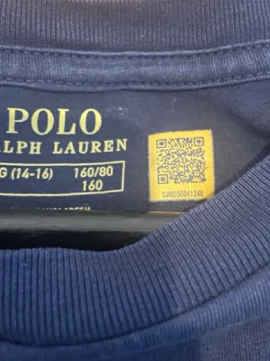 Mörkblå t-shirt från Polo Ralph Lauren - Snygg mörkblå t-shirt från Polo Ralph Lauren med klassisk röd broderad logga på bröstet. T-shirten har rund hals och korta ärmar, tillverkad i mjuk bomull som är skön mot huden. Perfekt för dig som gillar stilrena och ikoniska plagg.
