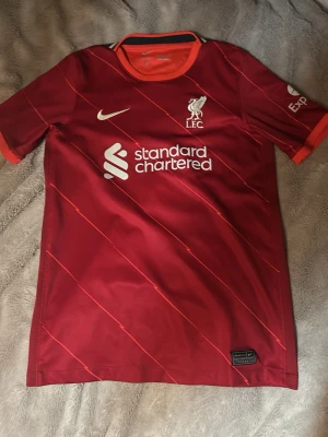 Liverpool FC Nike matchtröja röd 147 - 158 cm - Säljer en officiell Liverpool FC matchtröja från Nike. Storleken är gjort för personer som är cirka 147-158 cm lång. Tröjan är röd med tunna diagonala ränder och har vita logotyper samt sponsortryck. Tillverkad i lätt polyester med Dri-Fit teknologi för extra komfort. Perfekt för dig som älskar fotboll och Liverpool.