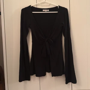 Svart omlottopp med knyt från Designby Si - Svart omlottopp från Design Si i storlek XS/S. Toppen har långärmade utsvängda ärmar, djup v-ringning och snygg knytning framtill. Materialet känns mjukt och stretchigt, perfekt för att skapa en cool och stilren look. Lite nopprig då den är använd men inget som syns direkt. 