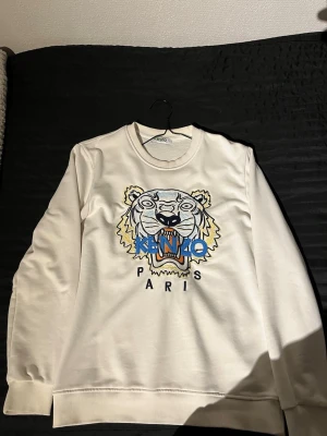 Vit sweatshirt från Kenzo - Säljer en vit sweatshirt från Kenzo med ikoniskt broderat tigermotiv och texten 'KENZO PARIS' på bröstet. Tröjan har rund hals, ribbade muddar och är långärmad. Perfekt statement-piece för dig som gillar streetwear och snygga detaljer.