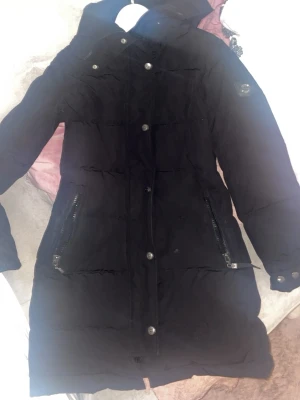 Svart lång dunjacka från Hollies - Säljer en svart dunjacka från Hollies Stockholm i storlek 34. Jackan är lång med huva, har snygga detaljer som silverfärgade knappar och dragkedjor vid fickorna. Den kommer med en fluffig päls på luvan, har bara inte inte med det i bilderna😊🥰