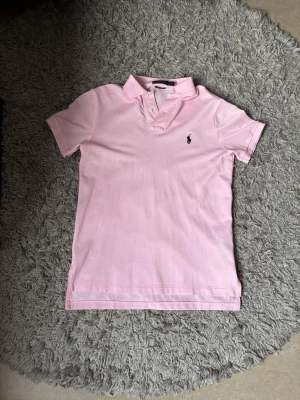 Rosa pikétröja från Ralph Lauren - Säljer en ljusrosa pikétröja från Ralph Lauren med klassisk krage och korta ärmar. Tröjan har två knappar vid halsen och den ikoniska broderade loggan i marinblått på bröstet. Perfekt för en avslappnad och stilren look.