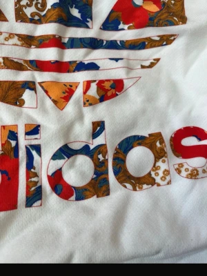 Vit croppad hoodie från Adidas - Säljer en vit croppad hoodie från Adidas med blommigt mönster i rött, blått och brunt på loggan och texten. Tröjan har långa ärmar med svarta ränder längs sidorna och huva med snörning. Perfekt för dig som gillar streetstyle och färgglada detaljer.