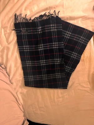 Mörkblå halsduk från Burberry - Mörkblå halsduk från Burberry i klassiskt rutigt mönster med gråa och röda toner. Halsduken är i 100% merinoull, perfekt för kallare väder! Pris kan diskuteras.