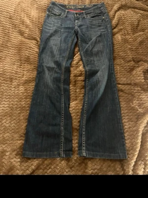 Blå bootcut jeans från Esprit - Säljer ett par blå jeans från Esprit med klassisk bootcut och låg midja. Jeansen har snygga detaljer med knappar på bakfickorna och kontrastsömmar. Materialet är slitstarkt denim i bomull och passformen är loose med breda benslut.