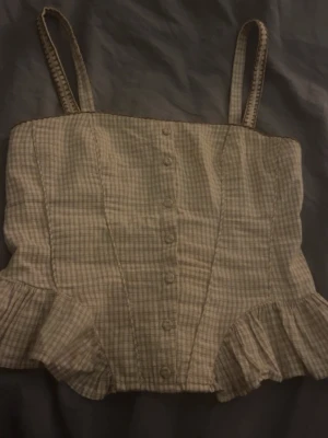 Beige rutig korsett med volang - Supersöt korsett-topp i beige med rutigt mönster och smala axelband. Toppen har knappar framtill, volangdetaljer nedtill och en smockad rygg för extra komfort. Materialet känns som bomull och toppen har även en dragkedja i sidan för enkel påklädning.