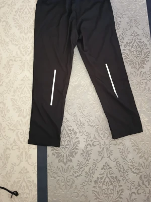 Svarta träningsbyxor med reflexdetaljer - Säljer ett par svarta träningsbyxor med smal passform och resår i midjan med snörning. Byxorna har två vertikala reflexdetaljer på framsidan av benen som gör dig synlig i mörkret. Perfekta för löpning eller gymmet.