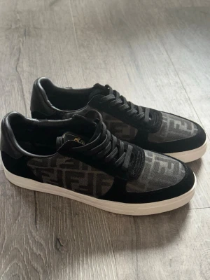 Svarta Fendi sneakers med mönster - Säljer ett par svarta Fendi sneakers med vit platt sula och snörning. Skorna har ett tydligt Fendi-mönster i grått och svart på sidan och ovandelen, samt detaljer i skinn och textil. Snygg och modern design med rund tå och klassisk Fendi-logga på insidan.
