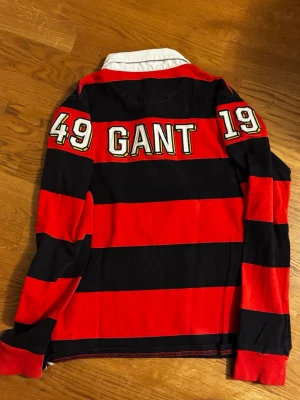 Röd och svart randig GANT skjorta - Snygg långärmad skjorta från GANT med breda röda och svarta ränder, vit krage och broderad G på bröstet. Skjortan har knappar vid halsen och tryck med siffror och logga på ryggen. Perfekt för dig som gillar sportig stil.