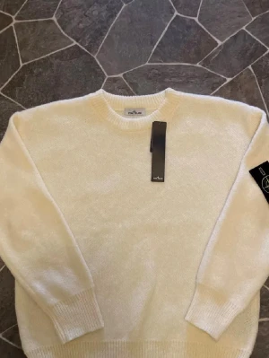 Vit stickad tröja från Stone Island - Säljer en fräsch vit stickad tröja från Stone Island med klassisk rund hals och ribbade muddar. Tröjan har den ikoniska Stone Island-patchen på vänster ärm och är tillverkad i mjukt material som passar perfekt för kyliga dagar.